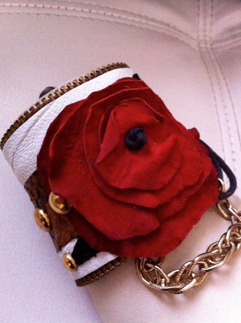 以下が含まれることがあります： 白と茶色の革製のブレスレット。大きな赤い布のバラと金のチェーンのアクセントが付いている。