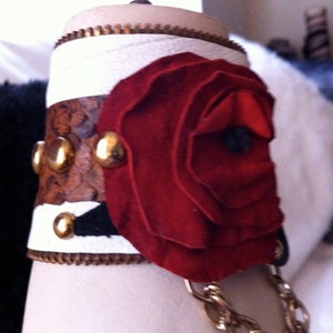 以下が含まれることがあります： ジッパー留めとゴールドチェーンが付いた白い革のブレスレット。ゴールドのアクセントがついた赤い布のバラで飾られています。