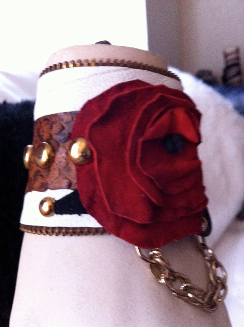 以下が含まれることがあります： ジッパー留めとゴールドチェーンが付いた白い革のブレスレット。ゴールドのアクセントがついた赤い布のバラで飾られています。