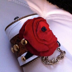 以下が含まれることがあります： 白と茶色の革製のカフブレスレット。大きな赤い布の花と金色のチェーンが付いています。花の中心は黒です。