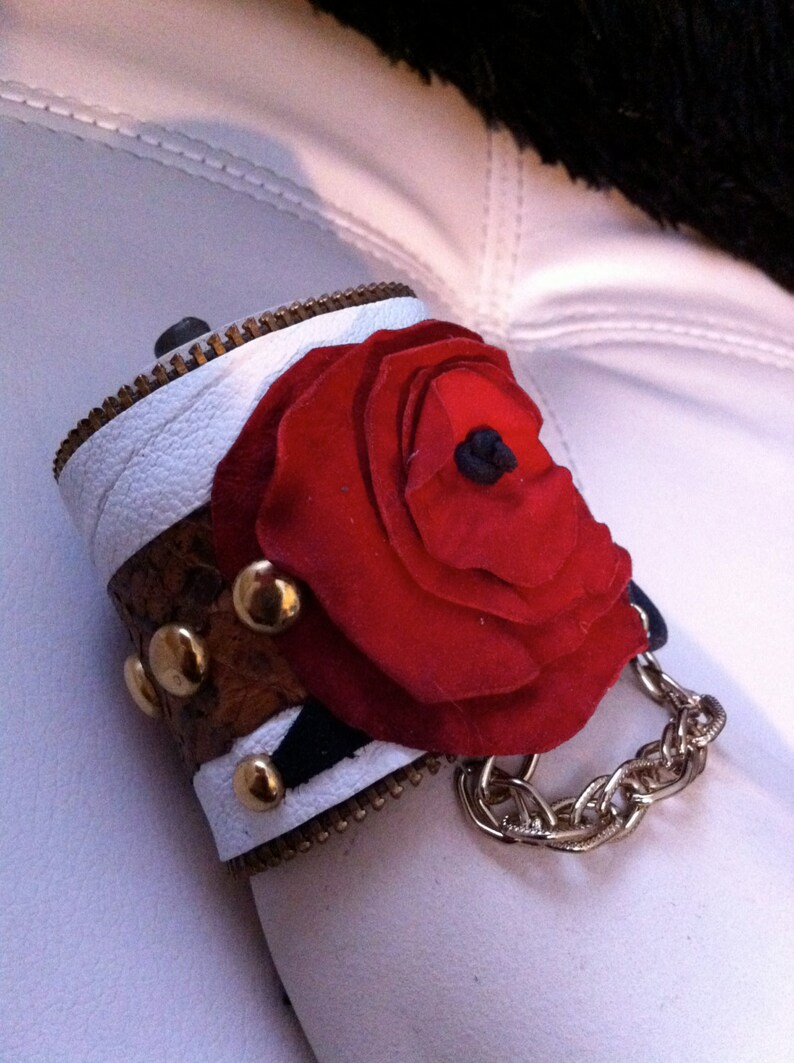 以下が含まれることがあります： 白と茶色の革製のカフブレスレット。大きな赤い布の花と金色のチェーンが付いています。花の中心は黒です。
