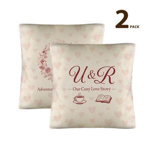 May include: Two decorative throw pillows with a cream background and a pattern of small pink hearts. One pillow features a floral wreath, the other has the text "U & R - Our Cozy Love Story" with a coffee cup and book illustration. The pillows are sold as a 2-pack.