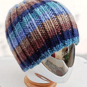 Peut inclure: Un bonnet tricoté à la main dans un mélange de couleurs bleues, marron et beige. Le bonnet a une texture côtelée et une couronne arrondie. Le chapeau est présenté sur une surface blanche.