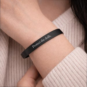 May include: A matte black cuff bracelet featuring the engraved white text "Protect the dolls." The bracelet has a smooth, solid band design with a clean finish, displaying the phrase clearly across the front surface of the metal accessory.