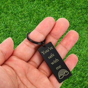 May include: A rectangular black metal keyring featuring a split ring and an engraved inscription that reads You're safe with me, accompanied by a small graphic of a rainbow with clouds at the base of the text.