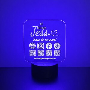 May include: A clear acrylic night light with the text "All Things Jess" and social media icons. The light has a black base and is illuminated with a blue glow. Includes QR codes for easy access to social media.
