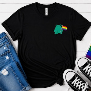 May include: Black t-shirt with a teal frog holding a rainbow flag. The shirt is styled with blue denim jeans and black and white trainers. A rainbow flag is also present. The t-shirt is a casual wear item.