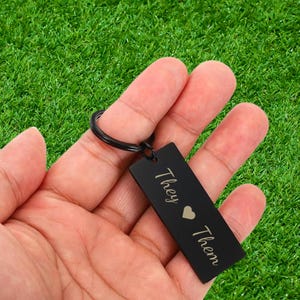 May include: A rectangular black metal keyring featuring a split ring and an engraved design. The text "They" is positioned above a small heart symbol, followed by the word "Them" below it, all written in a clean, light-coloured script font.