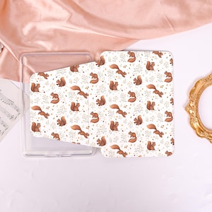 May include: A clear, rectangular phone case with a patterned cover. The cover features a repeating design of brown squirrels, yellow flowers, and green leaves on a white background. The case is open, revealing the interior.