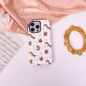May include: A white smartphone case featuring a repeating pattern of brown squirrels and small yellow and green floral sprigs. The case has a rectangular shape with rounded corners and a black camera cutout area at the top left corner.