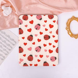 May include: A rectangular tablet case featuring a repeating pattern of red and pink ladybugs interspersed with red and pink hearts on a cream-colored background. The case has a tri-fold design with visible vertical creases across the surface.
