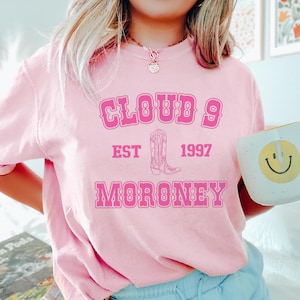 May include: Pink sweatshirt with the text "CLOUD 9 EST 1997 MORONEY" in a bubble font. A pink cowboy boot graphic is between "EST" and "1997". A white mug with a yellow smiley face is held in the right hand.