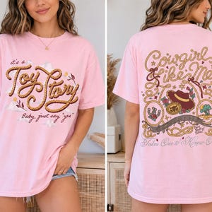 Op de afbeelding: Een lichtroze t-shirt met een voorzijde voorzien van touwachtige tekst "it's a Toy Story Baby, just say 'yes'" en een achterzijde met touwranden, een cowboyhoed en de tekst "Cowgirl like Me" en "Takes One to Know One."