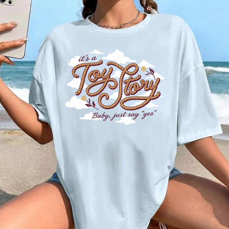 May include: A light blue short-sleeve t-shirt featuring a graphic print of white clouds and the text "it's a Toy Story Baby, just say "yes"" written in a brown rope-style font with small floral accents on the front center of the garment.