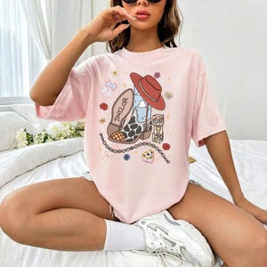 May include: A light pink t-shirt featuring a graphic print of a cowboy boot with a cow print pattern, a red cowboy hat, a stack of blocks with the letters T, S, and 13, a chain, flowers, and the word TAYLOR printed on the boot shaft.