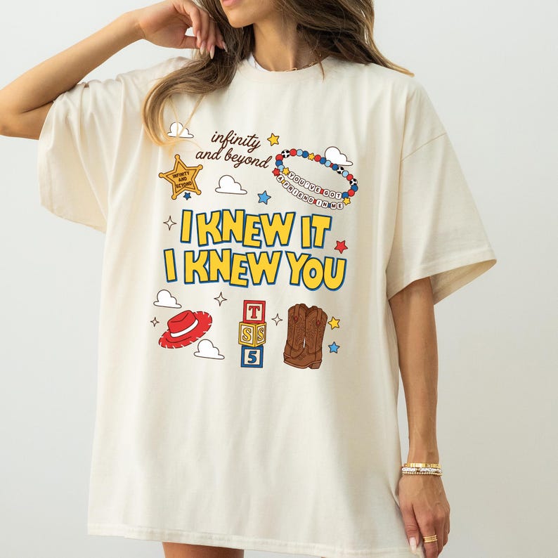 May include: An off-white short-sleeve t-shirt featuring a graphic print with the text "infinity and beyond", "I KNEW IT", "I KNEW YOU", and "YOU'VE GOT A FRIEND IN ME". The design includes a sheriff badge, a beaded bracelet, a red hat, alphabet blocks, and brown cowboy boots.