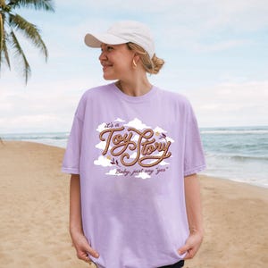May include: A light purple short-sleeve t-shirt featuring a graphic design of white clouds and brown rope-textured lettering that reads "it's a Toy Story" with the additional text "Baby, just say "yes"" below it, paired with a plain white baseball cap.