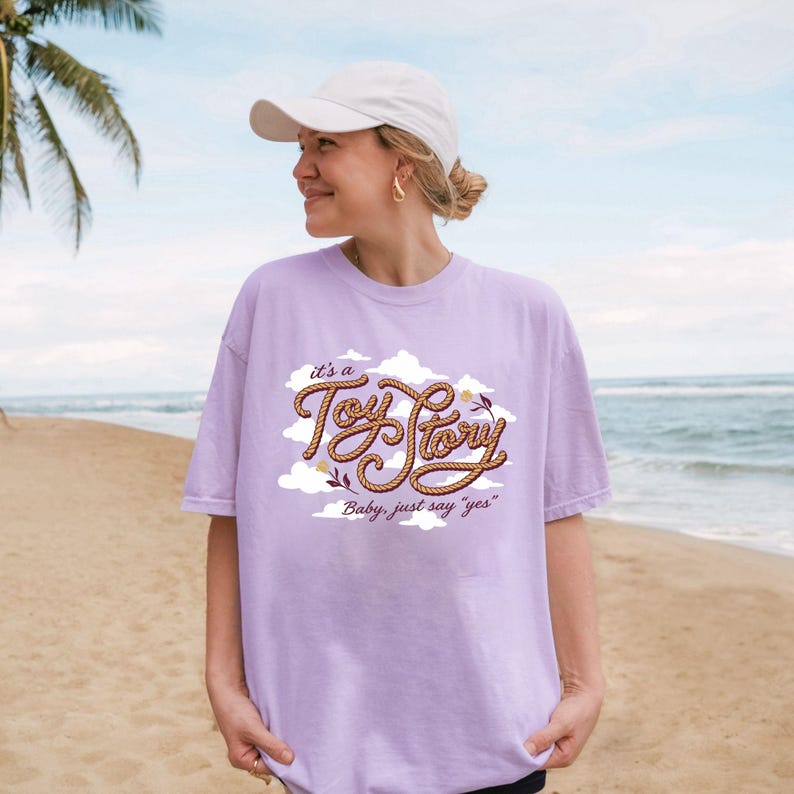 May include: A light purple short-sleeve t-shirt featuring a graphic design of white clouds and brown rope-textured lettering that reads "it's a Toy Story" with the additional text "Baby, just say "yes"" below it, paired with a plain white baseball cap.