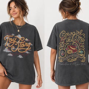 Op de afbeelding: Een antracietgrijs t-shirt met korte mouwen en een vooropdruk met de tekst Toy Story in een touwachtig lettertype boven wolken. De achterkant toont de tekst Cowgirl Like Me en Takes One to Know One met een illustratie van een hoed en cactus.