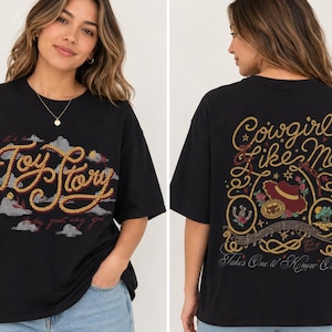 Op de afbeelding: Een zwart t-shirt met een vooropdruk van de tekst "Toy Story" in een touwachtig lettertype boven grijze wolken. De achterkant toont de tekst "Cowgirl like Me" en "Takes One to Know One" met illustraties van een hoed, hoefijzer en touw.