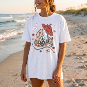 May include: A white short-sleeve t-shirt featuring a graphic print of a cow-print cowboy boot with the name Taylor, a red cowboy hat, a chain, and stacked blocks with the letters T, S, and the number 13, surrounded by small floral and star icons.