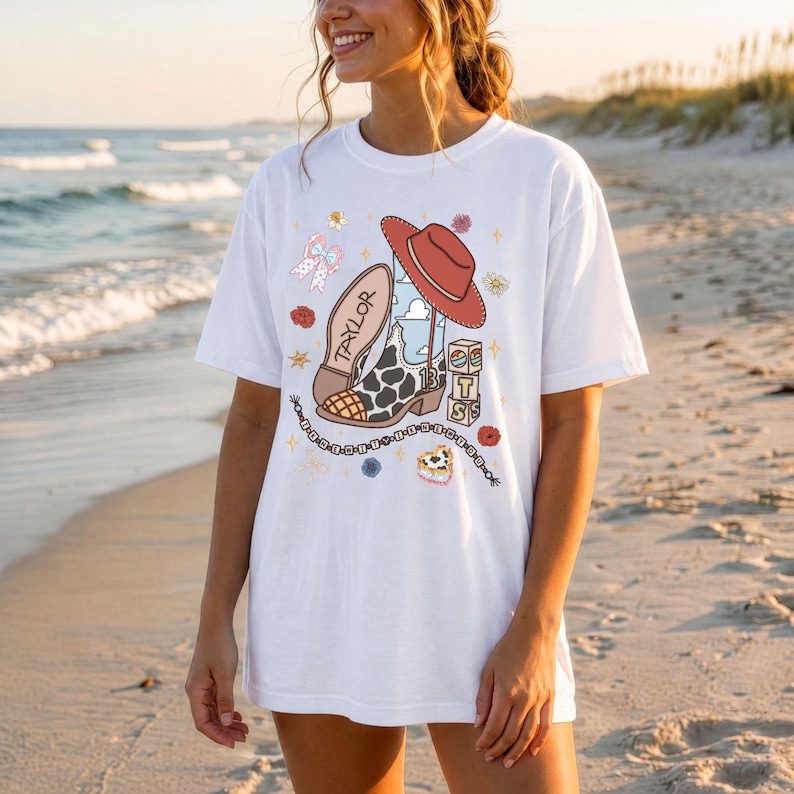 May include: A white short-sleeve t-shirt featuring a graphic print of a cow-print cowboy boot with the name Taylor, a red cowboy hat, a chain, and stacked blocks with the letters T, S, and the number 13, surrounded by small floral and star icons.