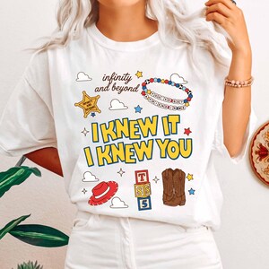 May include: A white t-shirt featuring a graphic print with the text "infinity and beyond", "YOU'VE GOT A FRIEND IN ME", and "I KNEW IT I KNEW YOU". The design includes a yellow sheriff star, a beaded bracelet, a red hat, alphabet blocks, and boots.