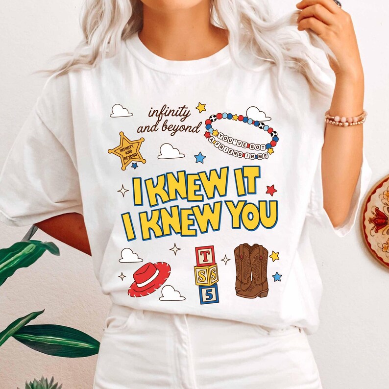 May include: A white t-shirt featuring a graphic print with the text "infinity and beyond", "YOU'VE GOT A FRIEND IN ME", and "I KNEW IT I KNEW YOU". The design includes a yellow sheriff star, a beaded bracelet, a red hat, alphabet blocks, and boots.