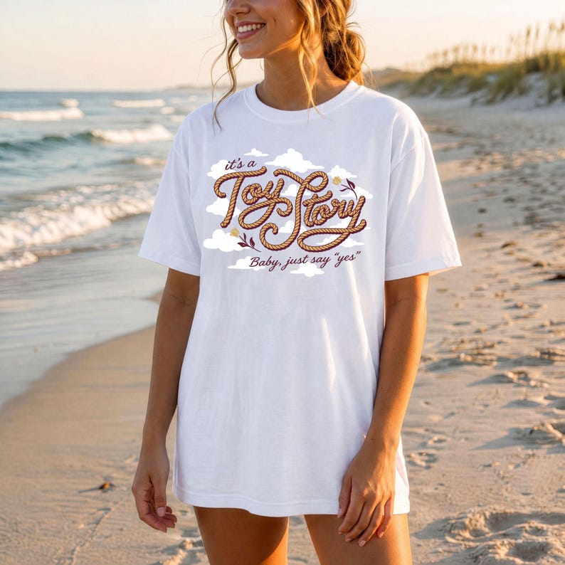 May include: A white short-sleeved t-shirt featuring a graphic design of brown rope shaped into the text "it's a Toy Story" surrounded by white clouds and small flowers, with the additional text "Baby, just say "yes"" printed below in brown.