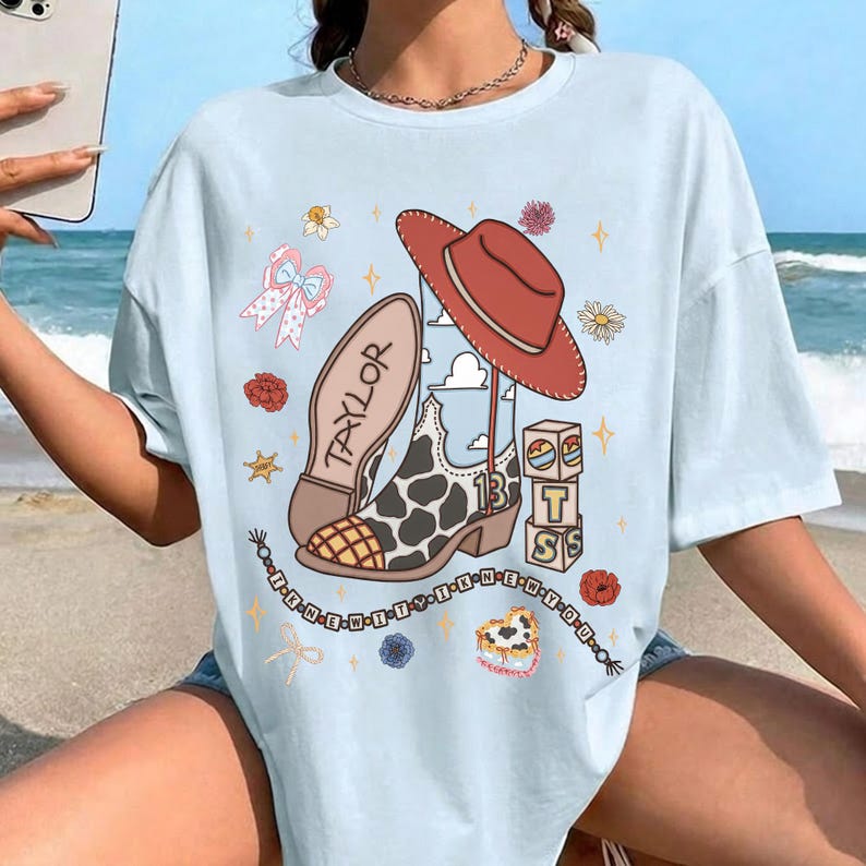 May include: A light blue t-shirt featuring a graphic print of a cow-print cowboy boot, a red cowboy hat, blocks with the letters T, S, and the number 13, a bow, flowers, and a beaded string with the text I KNEW YOU WERE TROUBLE.
