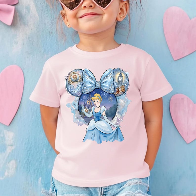 May include: A light pink t-shirt featuring a Cinderella graphic. The graphic is a blue bow with Cinderella's image in the center. The design includes a clock, a carriage, and other elements from the movie. The shirt is for children.