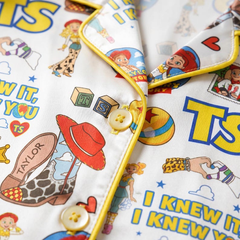 May include: A white pajama top with yellow piping and a repeating pattern of Toy Story characters, cowboy boots, alphabet blocks, and hearts. The fabric features the text I KNEW IT, I KNEW YOU, and TAYLOR printed in blue and yellow across the surface.