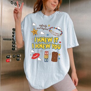 May include: A light blue t-shirt featuring a graphic print with the text "infinity and beyond", "I KNEW IT I KNEW YOU", and "YOU'VE GOT A FRIEND IN ME". The design includes a sheriff star, a beaded bracelet, clouds, a red hat, alphabet blocks, and boots.