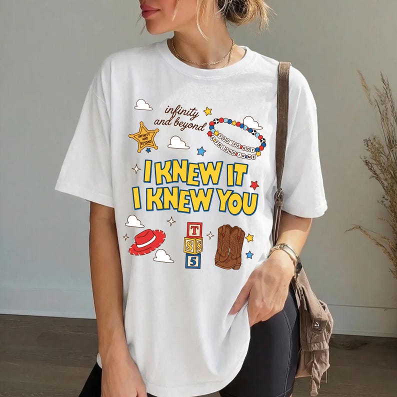 May include: A white short-sleeve t-shirt featuring a graphic print with the text "infinity and beyond," "YOU'VE GOT A FRIEND IN ME," "I KNEW IT," and "I KNEW YOU." The design includes a yellow sheriff badge, a red hat, alphabet blocks, and boots.