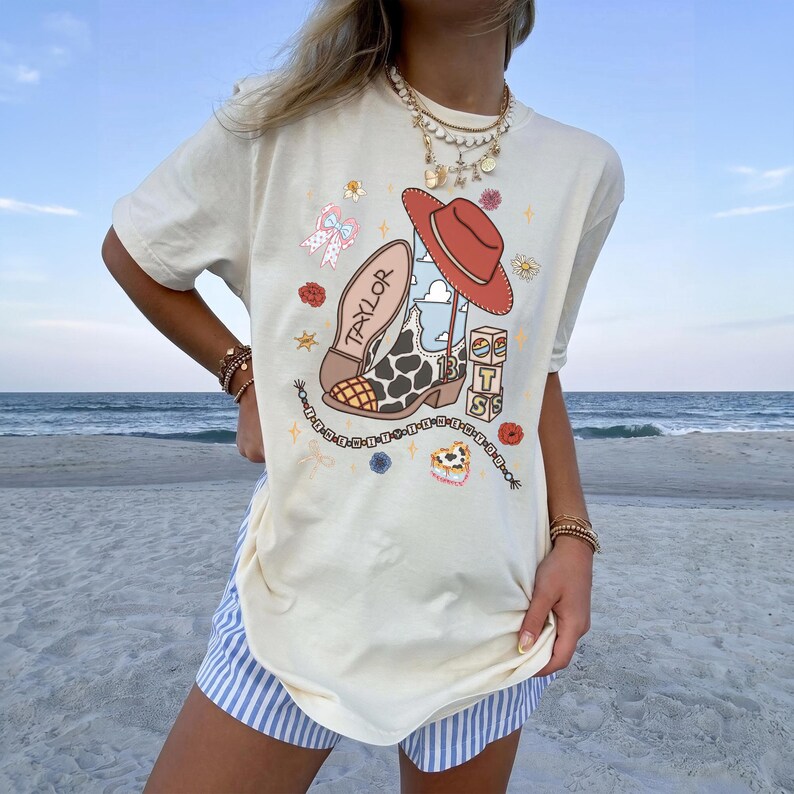 May include: A cream-coloured short-sleeve t-shirt featuring a graphic print of a cowboy boot with a cow print pattern, a red hat, a bow, and blocks with the letters T, S, and the number 13. The word TAYLOR is printed on the boot.