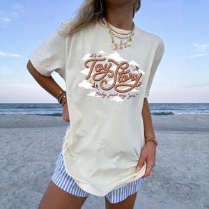 May include: A cream-colored short-sleeve t-shirt featuring a graphic design of brown rope-style lettering that reads "it's a Toy Story" and "Baby, just say "yes"" surrounded by white clouds and small floral accents on the front center.