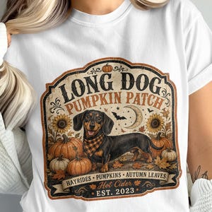 May include: A white t-shirt featuring a graphic of a black and tan dachshund wearing a checkered scarf. The design includes pumpkins, sunflowers, a crescent moon, bats, and the text LONG DOG PUMPKIN PATCH, HAYRIDES, AUTUMN LEAVES, Hot Cider, EST. 2023.