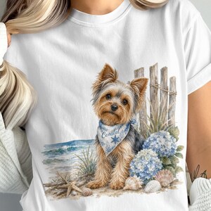 Op de afbeelding: Een wit t-shirt met een grafische illustratie van een Yorkshire Terriër die een blauwe bandana draagt met het woord Coastal erop gedrukt. De hond staat op een zandstrand met een houten hek, blauwe hortensia's en verschillende zeeschelpen.