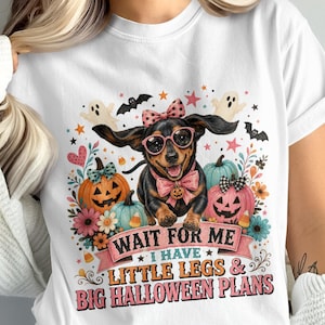 Puede incluir: Una camiseta blanca con un gráfico de un perro salchicha con gafas rosas y un lazo, rodeado de calabazas, fantasmas y flores. El texto dice: WAIT FOR ME I HAVE LITTLE LEGS & BIG HALLOWEEN PLANS en letras de color rosa y negro.