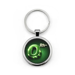 May include: A circular, silver-coloured keyring with a green and black pendant. The pendant displays a glowing green letter "O" with the letters "Rh-" in silver. The keyring is set against a white backdrop.