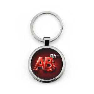 May include: A circular keyring with a silver-coloured metal frame. The centre displays the letters "AB" in a 3D, metallic style, and "Rh-" above, set against a red background. The keyring has a silver key ring.