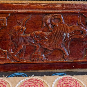 May include: A rectangular, dark brown wooden carving with detailed relief of mythical creatures. Above it, a decorative wooden piece with a blue center and a small red felt animal. Below, a beige fabric with embroidered pink roses.