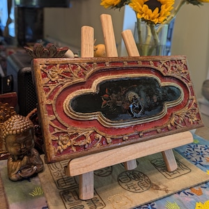 May include: A rectangular wooden decorative panel featuring intricate gold and red floral carvings surrounding a central black oval inlay with a small metal ring pull. The panel is displayed on a light-colored wooden tripod easel stand.