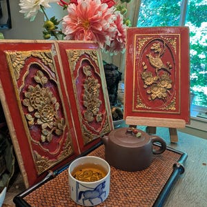 May include: Three rectangular red and gold wooden wall panels featuring carved floral and bird motifs. A brown clay teapot and a white ceramic tea cup with a blue horse illustration sit on a rectangular woven tray with black edges.