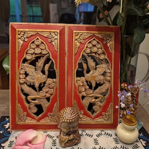 May include: Two red wooden panels with gold relief carvings of birds and foliage. A small stone Buddha statue sits in front of the panels. A vase of flowers and a small decorative vase are also present.
