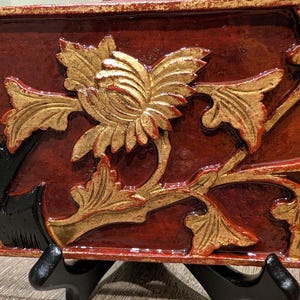 May include: A rectangular, ornate, wooden panel with a floral design. The panel features a deep red background with gold-colored floral and leaf details. The panel is supported by a black stand.