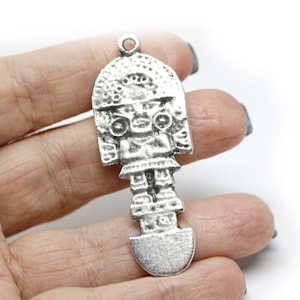 May include: Silver pendant featuring a detailed depiction of a stylized human figure with a headdress. The figure is standing with its arms at its sides and has a rounded base.
