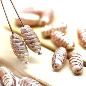 May include: A close-up of a collection of oval-shaped glass beads with a metallic rose gold finish. The beads have a swirl pattern and are strung on a wire.