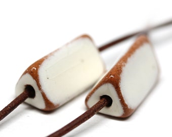 May include: Two white ceramic beads with brown accents are strung on a brown leather cord. The beads are triangular in shape and have a hole in the centre for threading.