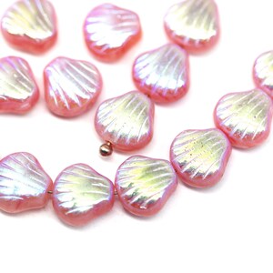 May include: A collection of pink, iridescent shell-shaped beads. Each bead has a textured, fan-like pattern and a pearlescent sheen. The beads are strung together, suggesting they are for jewelry making or crafting.